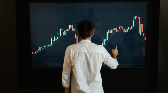 Adult male reviewing stock market data on a large display screen indoors.
